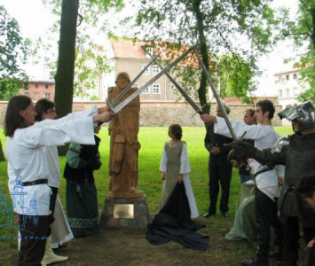 grupa przebranych młodych ludzi krzyżuje nad głowami miecze obok drewnianego pomnika